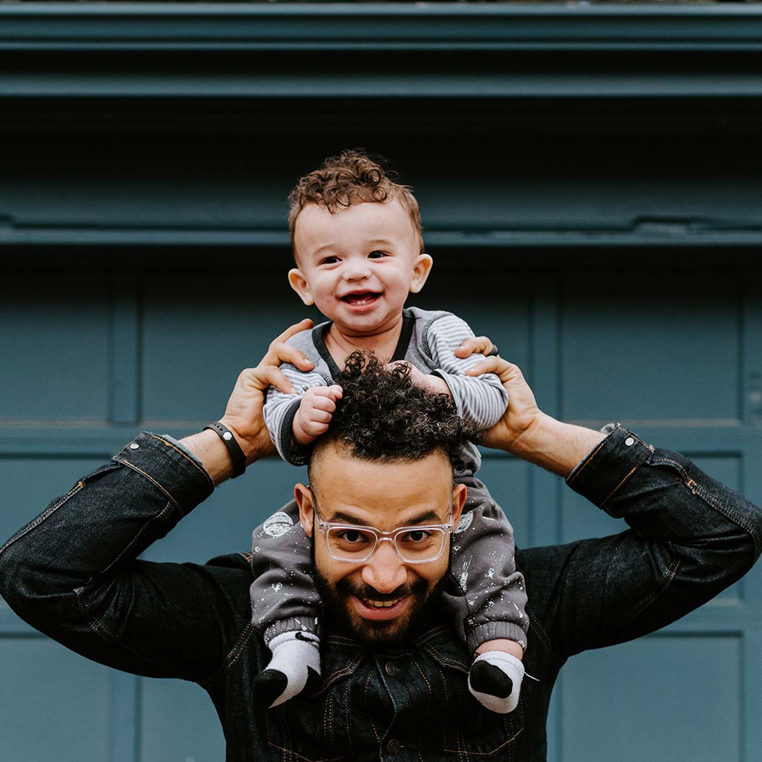 Hopeful and playful BIPOC parent wearing glasses with their child on their shoulders
