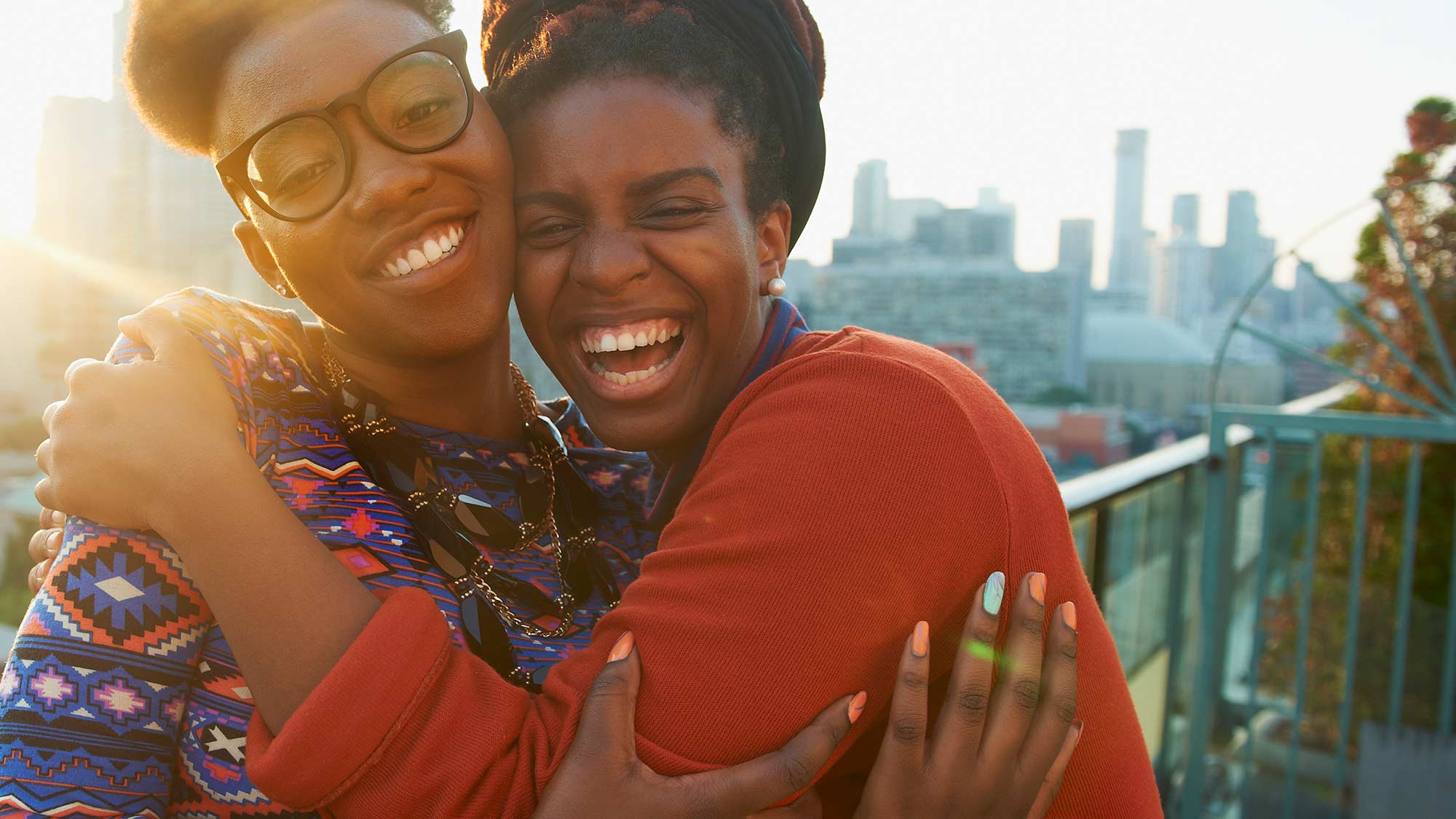 BIPOC women hugging and smiling on city rooftop at sunset